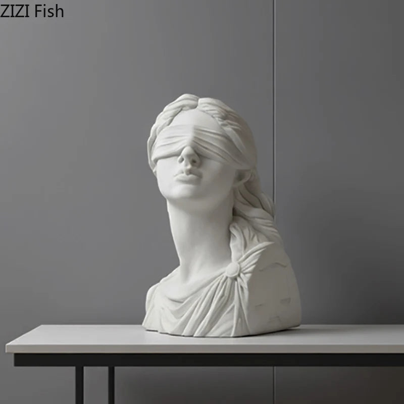 White marble bust of a woman with blindfold on a gray surface against a gray wall.