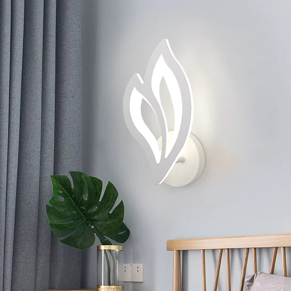 Modern wall sconce with leaf design on a light gray wall, next to a plant and wooden bed.