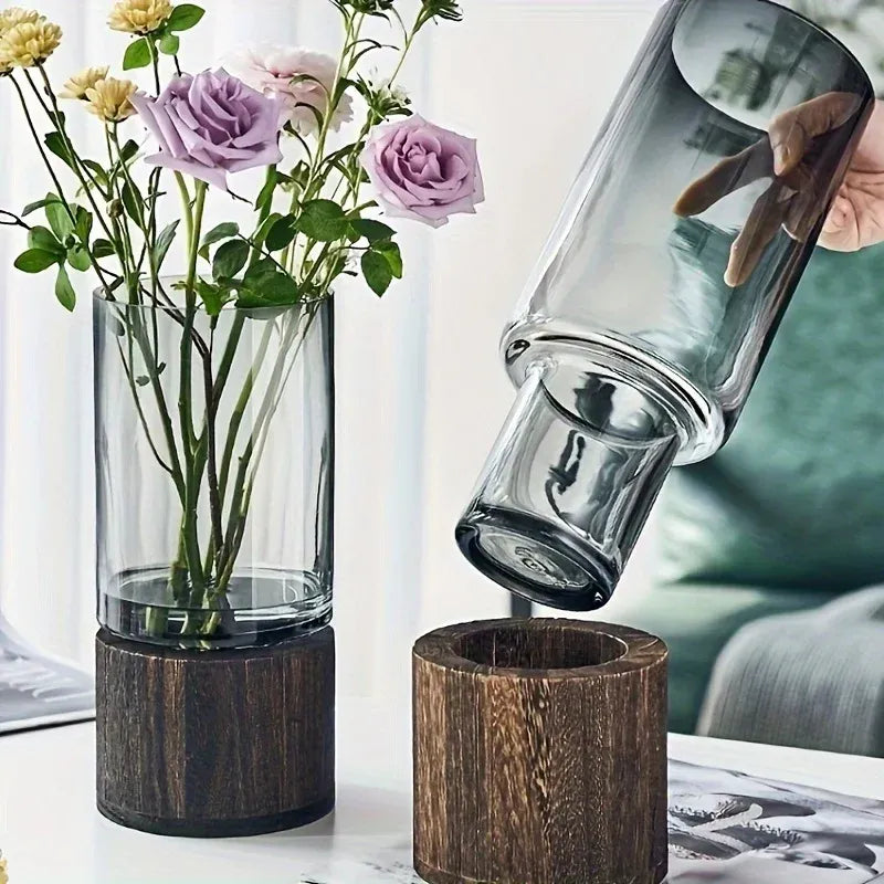 A glass carafe is being tilted over a wooden funnel with flowers in a vase on a table.