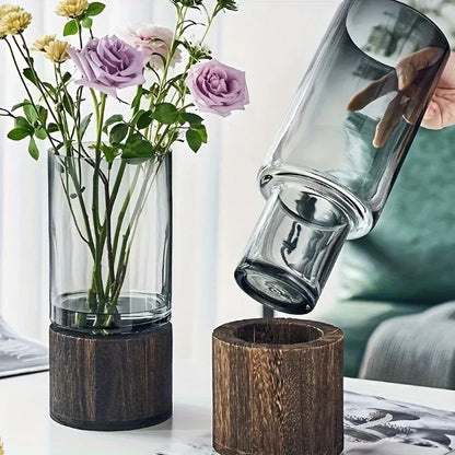 A glass carafe is being tilted over a wooden funnel with flowers in a vase on a table.
