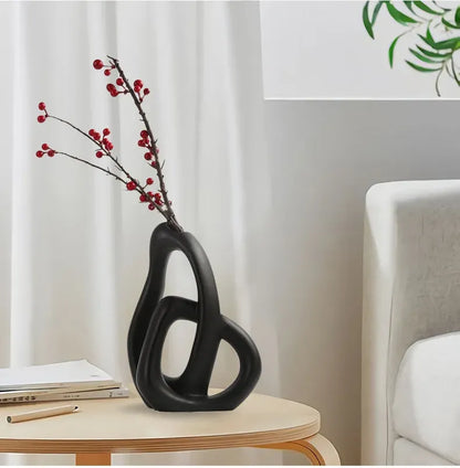Black abstract vase with red branches on a table in a room with a white curtain and chair.