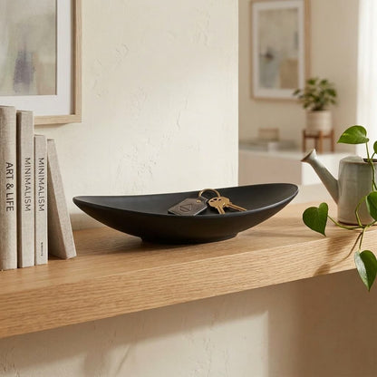 Black dish on a wooden shelf with keys, books, and a teapot in a home setting.