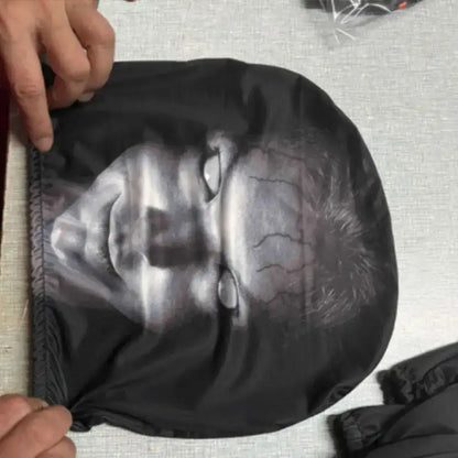Black horror covers with a printed face design held by a person on a textured surface.