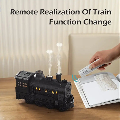 Black train-shaped device with steam effect on a wooden surface, held by a hand with a remote control.