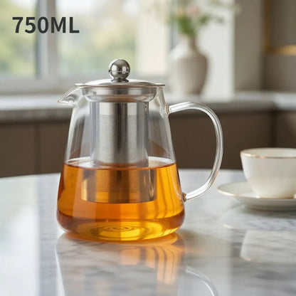 Glass teapot with metal lid and handle filled with tea on a marble surface.