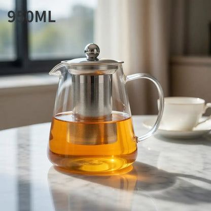 Glass teapot with metal infuser filled with tea on a marble surface, with a cup and saucer in the background.