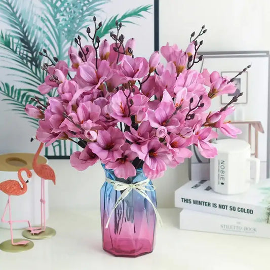 Bouquet of pink flowers in a gradient vase on a white surface with decorative items in the background.