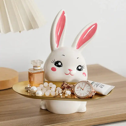 Ceramic bunny figurine with a gold tray holding jewelry and a bottle of perfume on a wooden surface.