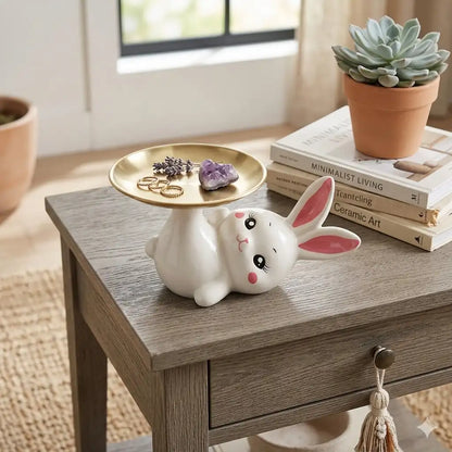 Ceramic rabbit with a small round table on a wooden surface, with books and a plant in the background.