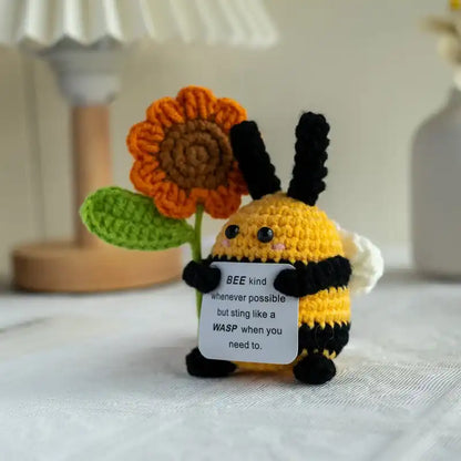 Crocheted bee toy with a flower and a humorous tag on a light surface.
