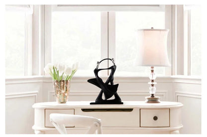 Decorative black sculpture on a white console table with a lamp and flowers in a room with large windows.