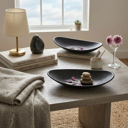Decorative setting with black bowls, books, a lamp, and flowers on a wooden table.