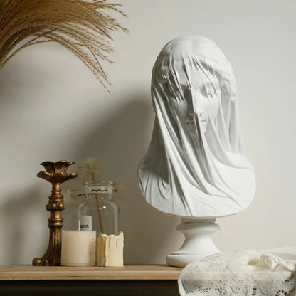 Decorative bust with a white cloth draped over it on a wooden surface with candles and a jar.