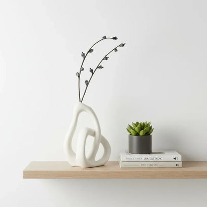 Decorative vase with branches, small plant, and books on a wooden shelf against a white wall.