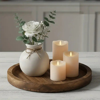 Decorative setup with a white vase holding flowers and three candles on a wooden tray.