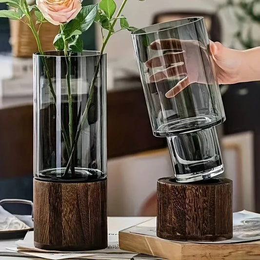 Glass vase with wooden base holding flowers, and a hand lifting another glass vase.