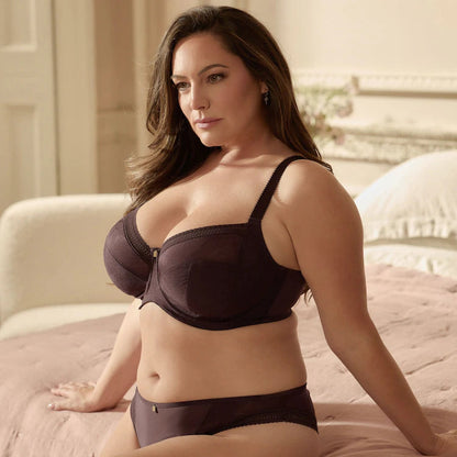 A woman wearing a dark plum purple bra and matching underwear sits on a bed with pink bedding in a softly lit bedroom.