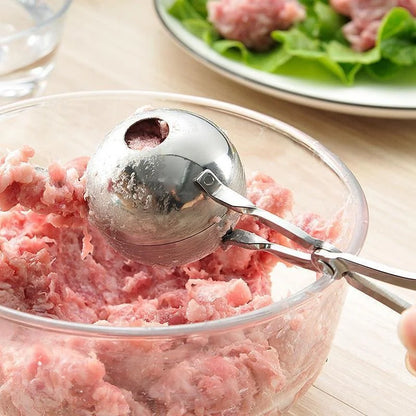 Metallic meatball maker in a bowl of ground meat with a plate of raw meat and salad in the background.