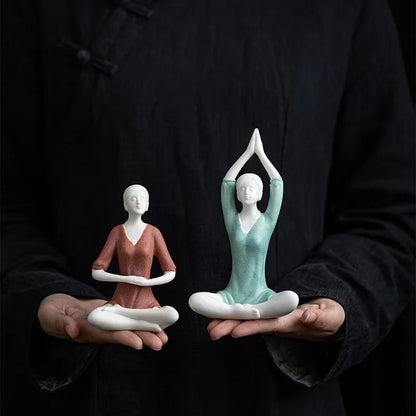 Two ceramic figures in yoga poses held in hands against a black background