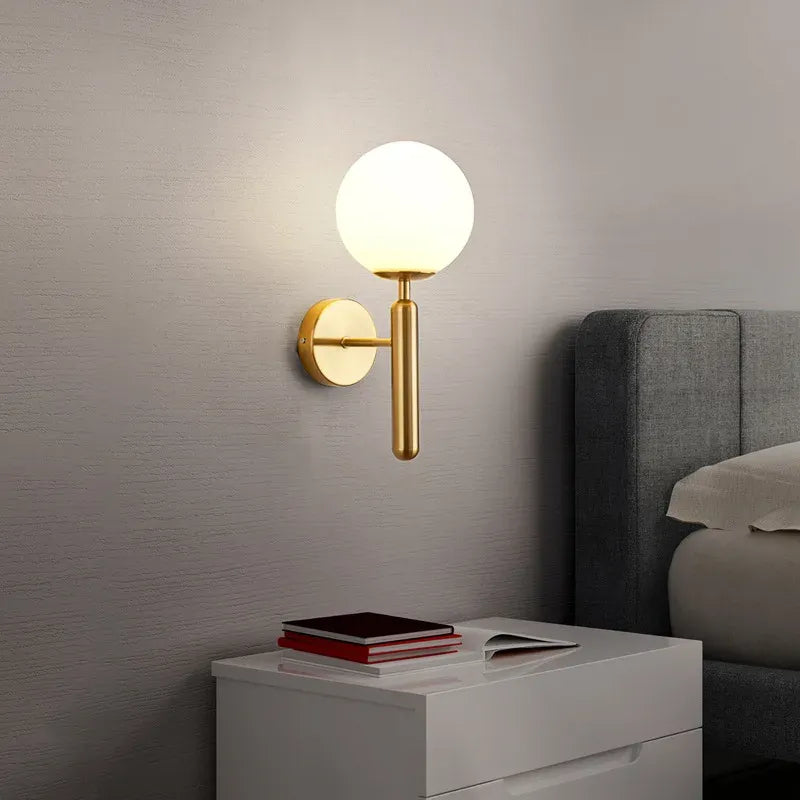 Modern wall sconce with a round light fixture on a gray wall next to a bed.