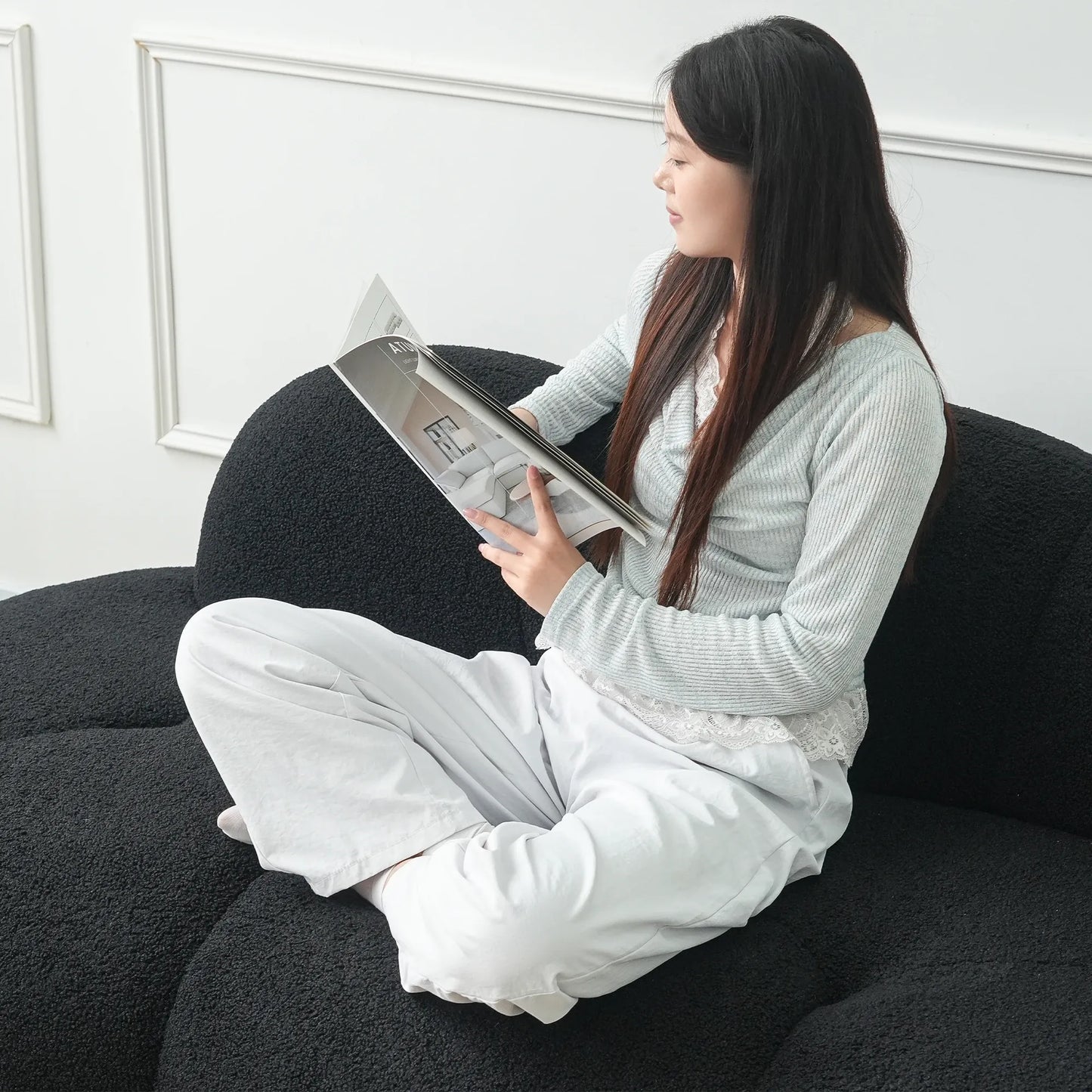 Woman sitting on a black sofa reading a magazine in a minimalistic room.