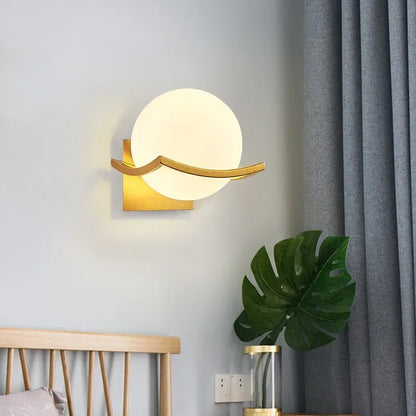 Modern wall lamp with gold frame and white globe on a gray wall next to a plant and bed.
