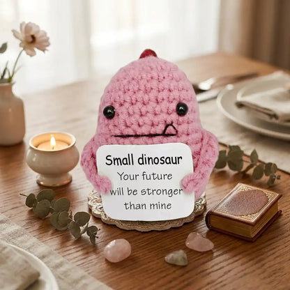 Pink crochet dinosaur with a message card on a table with a candle and flowers.