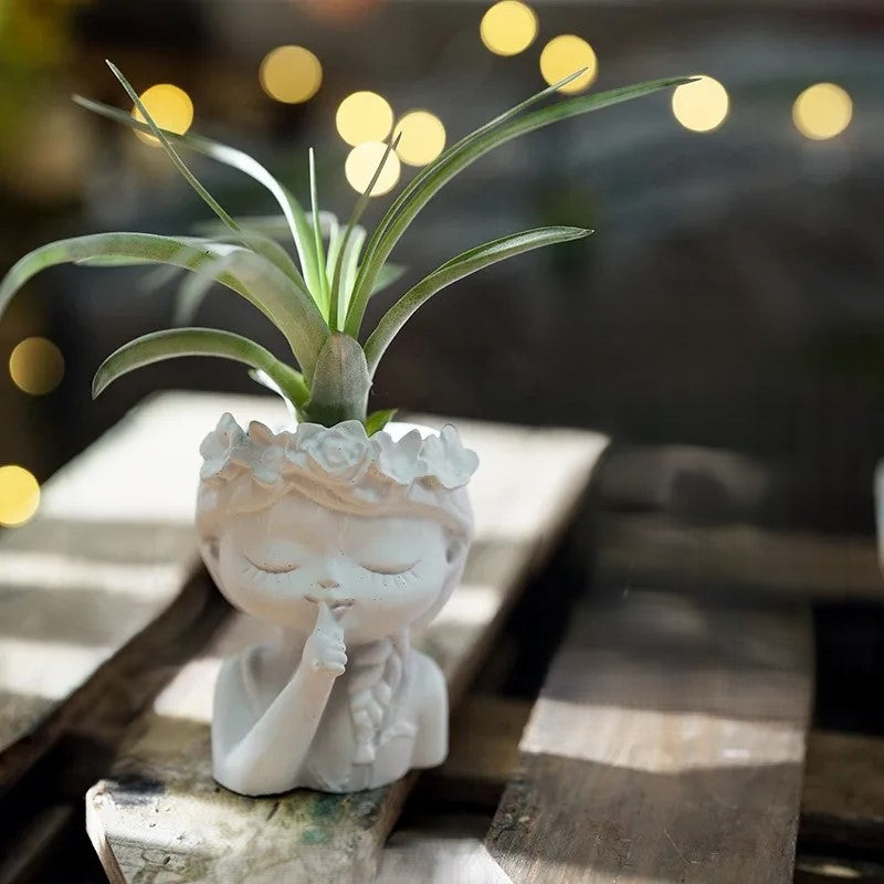 White statue of a person with a plant on a wooden surface with blurred lights in the background