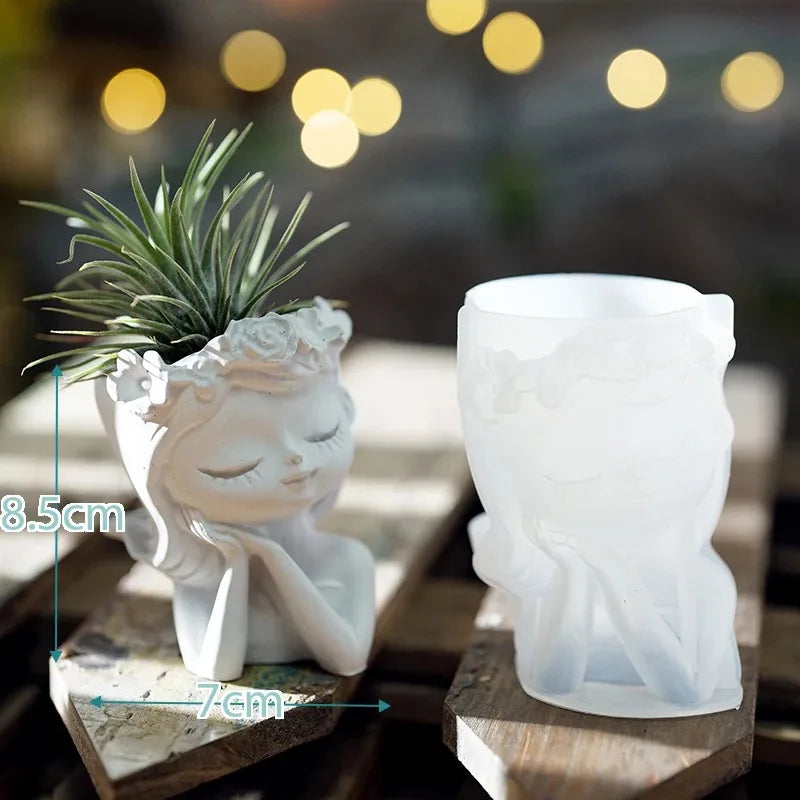 White ceramic planter shaped like a face with a plant on top, next to a plain white planter, on a wooden surface with blurred lights in the background.