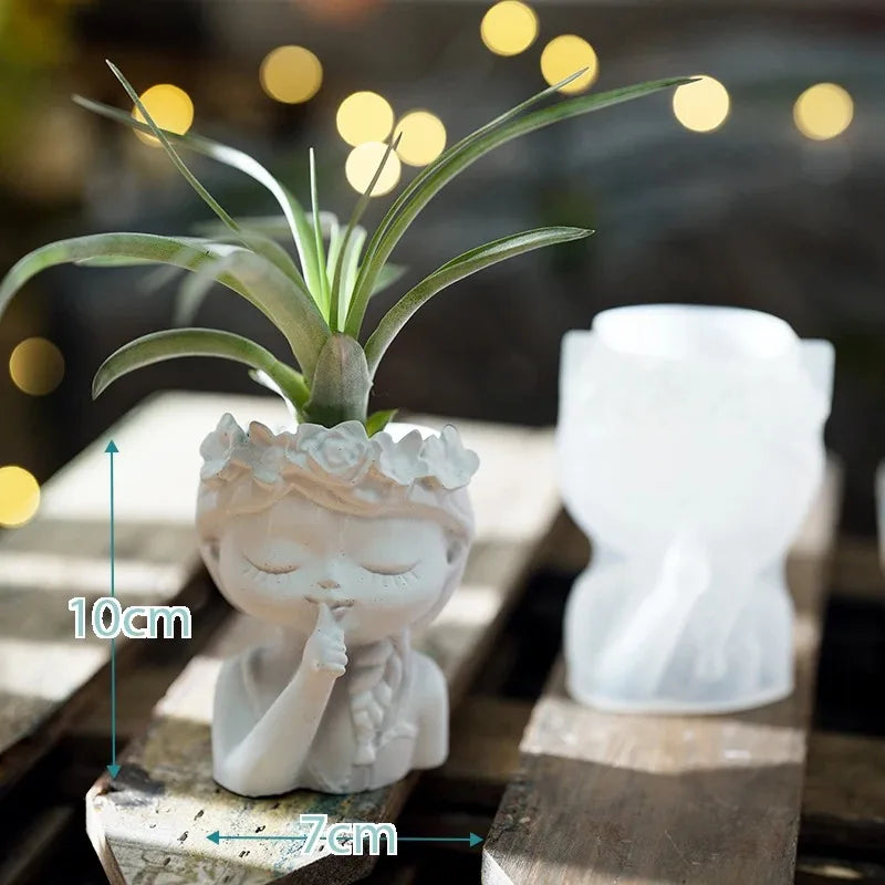 Small white statue with a plant on top, placed on a wooden surface with blurred lights in the background.
