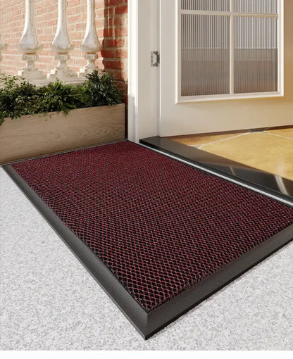 Red doormat with black border on a concrete floor outside a building entrance.