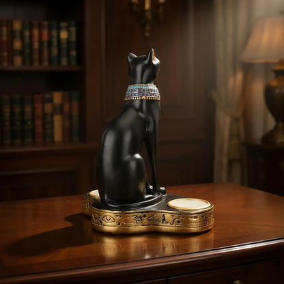 Statue of a black cat with a colorful collar on a wooden table in a room with books and a lamp.