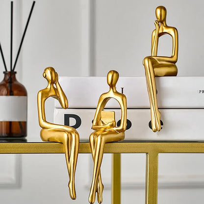Three gold-colored sitting figures on a shelf with a minimal background