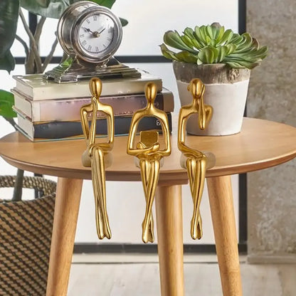 Three gold decorative figures on a wooden table with a clock and plant in the background.