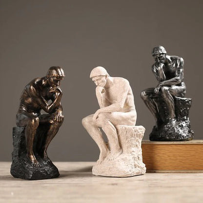 Three statues of 'The Thinker' in different materials and colors on a wooden surface with a gray background.