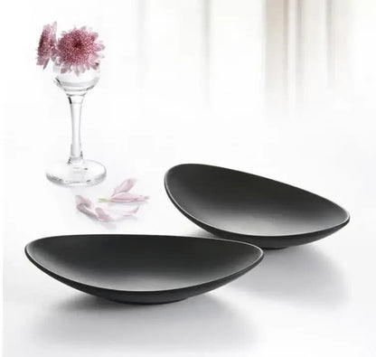 Two black ceramic bowls on a white surface with a glass and flowers in the background.