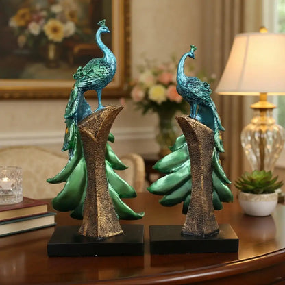 Two decorative peacock statues on a table with a lamp and books in the background.