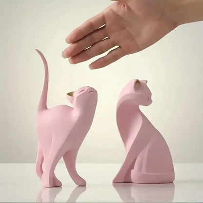 Two pink ceramic cat figurines on a light surface with a hand reaching towards them.