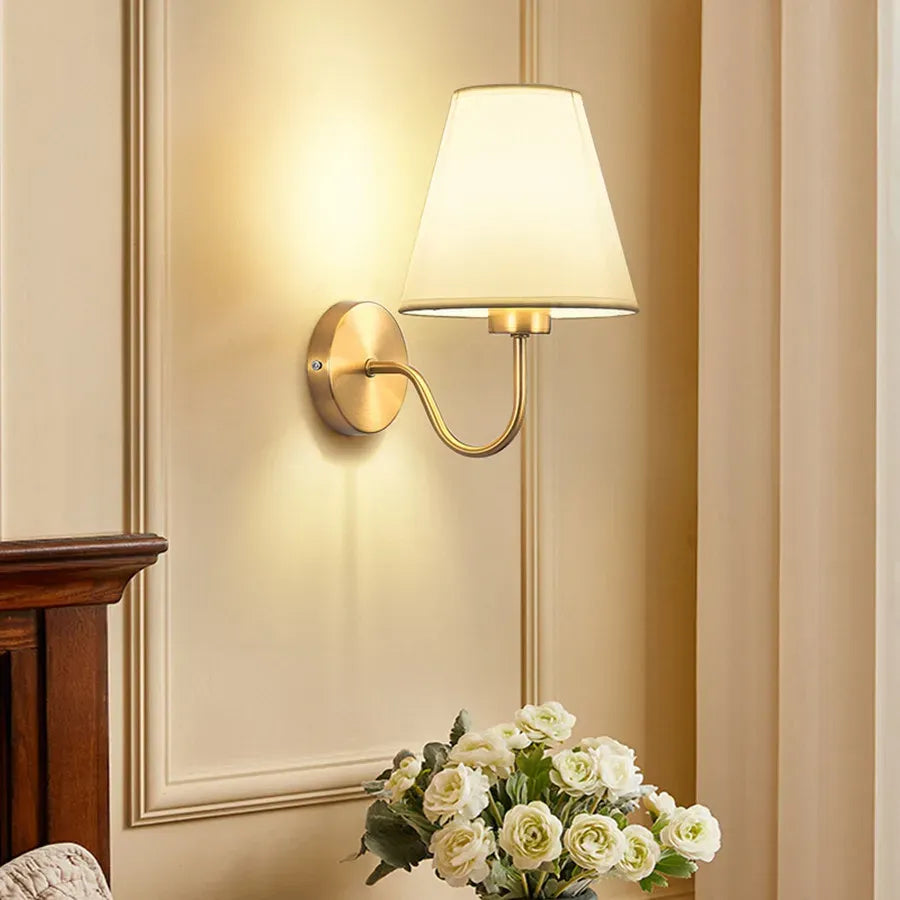 Wall-mounted lamp with a white shade on a beige wall, next to a wooden piece of furniture and a vase of flowers.