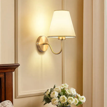 Wall-mounted lamp with a white shade on a beige wall, next to a wooden piece of furniture and a vase of flowers.