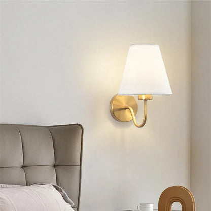 Wall-mounted lamp with a white shade and gold fixture on a neutral wall next to a bed.