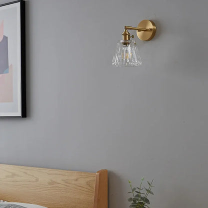 Wall-mounted lamp with clear glass shade on a gray wall above a wooden headboard.