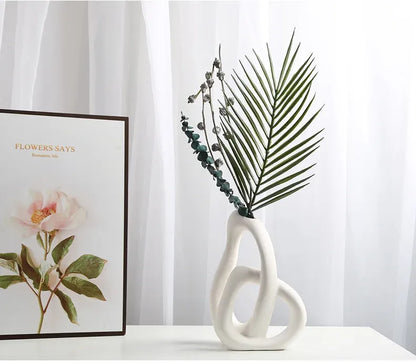 White vase with decorative plant on a white surface, with a floral-themed book in the background.