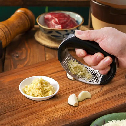 Hand using a garlic press on a wooden table with minced garlic and whole garlic cloves.