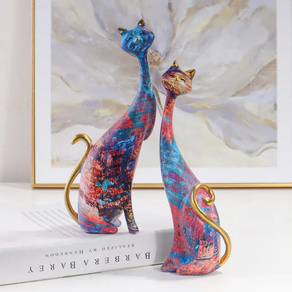 Colorful cat figurines on a surface with a framed abstract painting in the background.