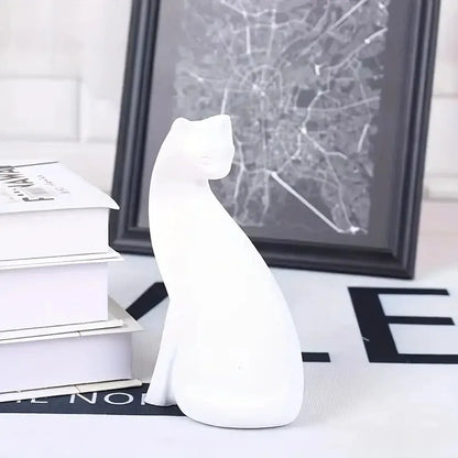 White ceramic cat figurine on a surface with books and a framed picture in the background
