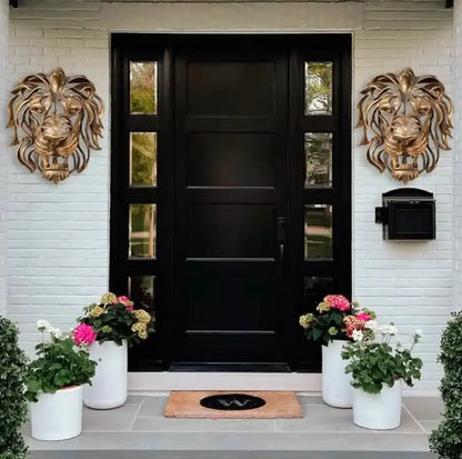 Black front door with decorative lion heads on a white brick wall, featuring potted plants and a welcome mat.