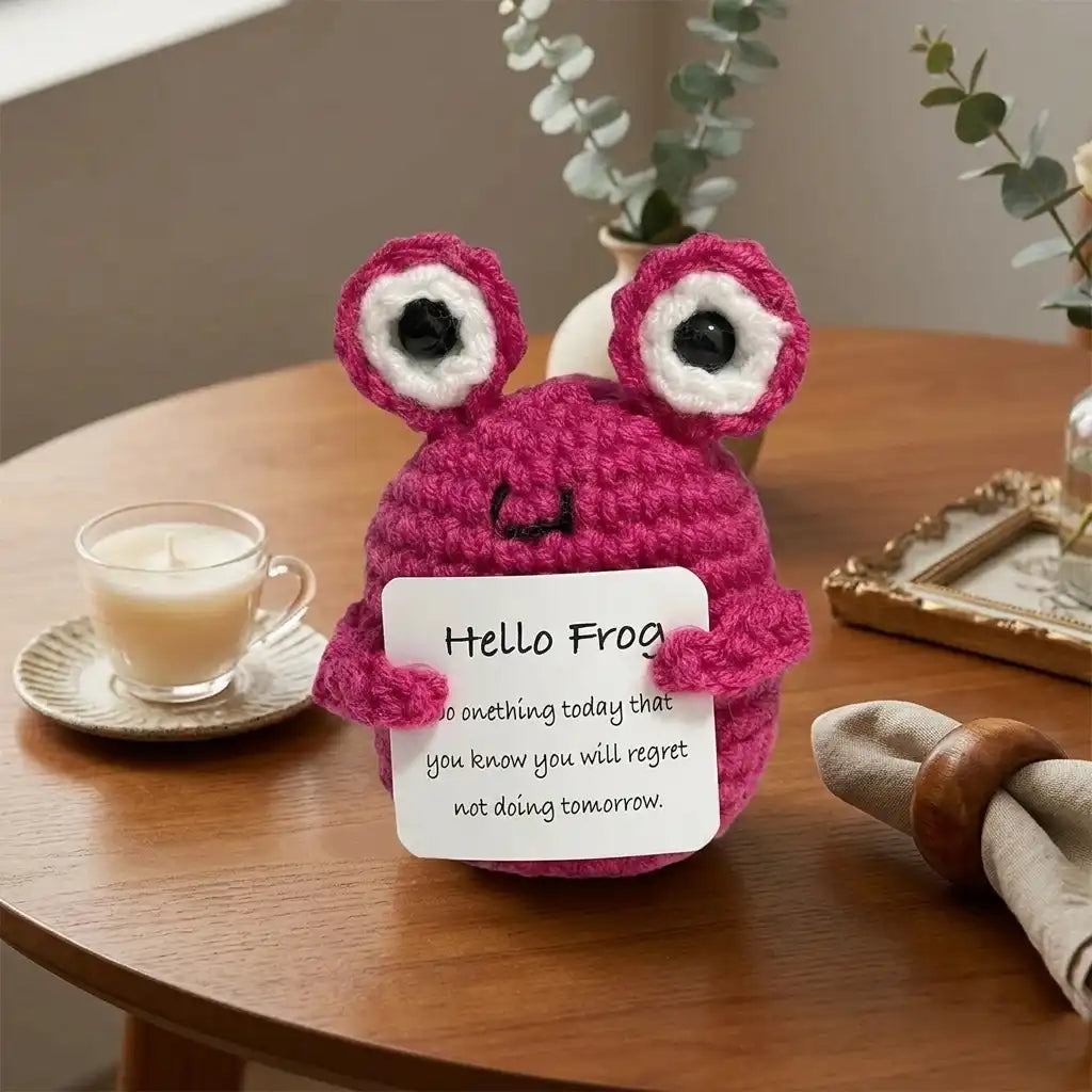Crocheted pink frog toy holding a sign on a wooden table with a cup of coffee and plants in the background.