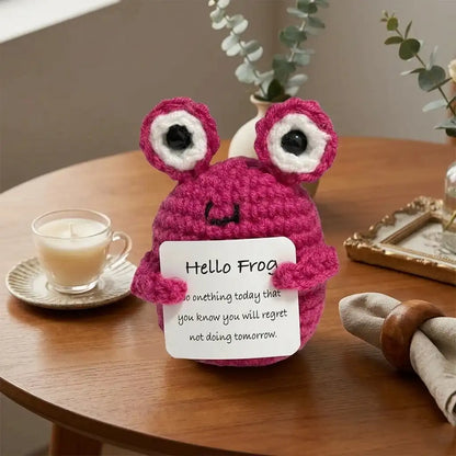 Crocheted pink frog toy holding a sign on a wooden table with a cup of coffee and plants in the background.