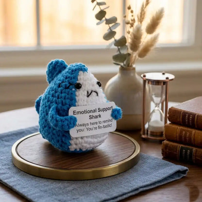 Crocheted blue and white shark plush toy holding a sign on a wooden coaster with books and decor in the background.
